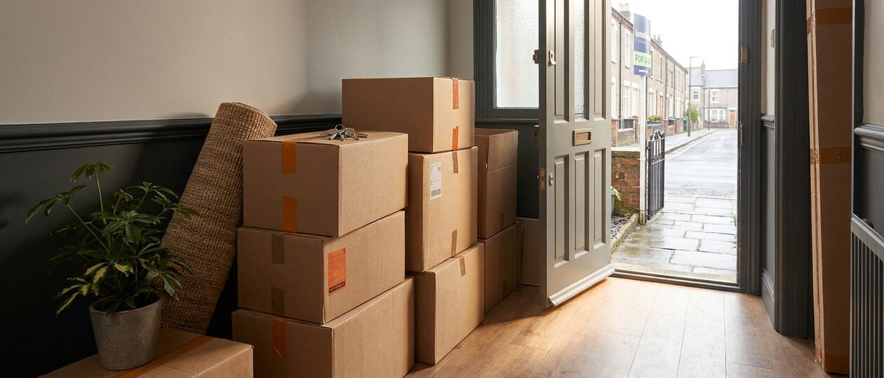 Moving boxes stacked in the hallway of a terraced house, the front door open onto a street with a For Sale sign